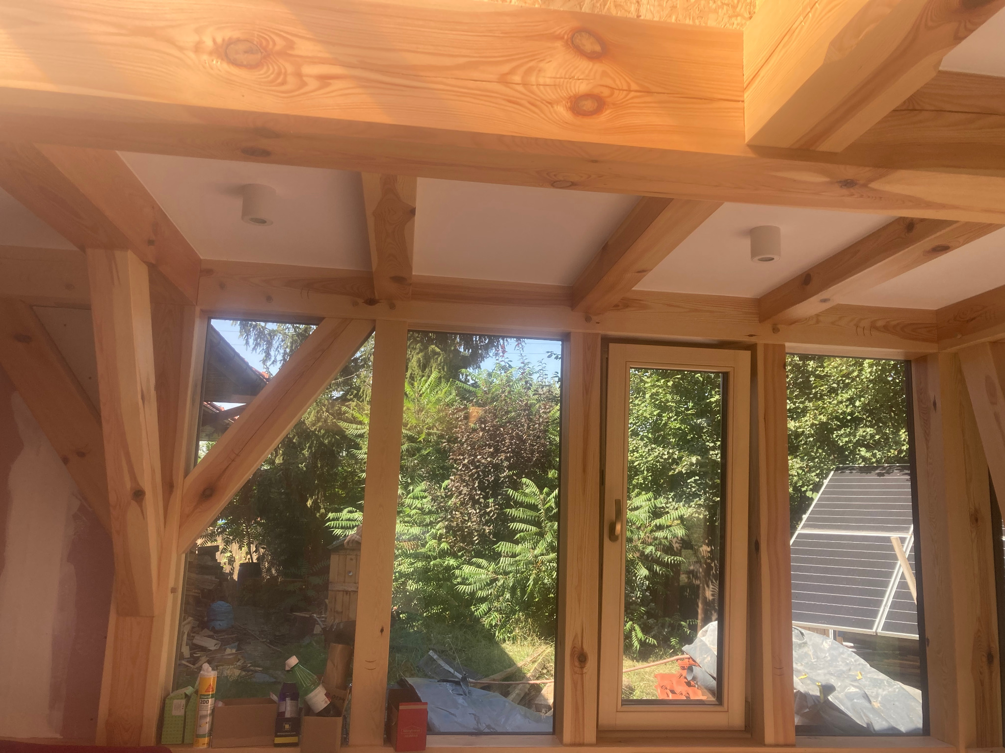 Wnętrze w trakcie wykończenia z drewnianą konstrukcją stropu i ramami okiennymi. Widoczne elementy instalacji oświetleniowej i widok na zewnątrz przez okna.