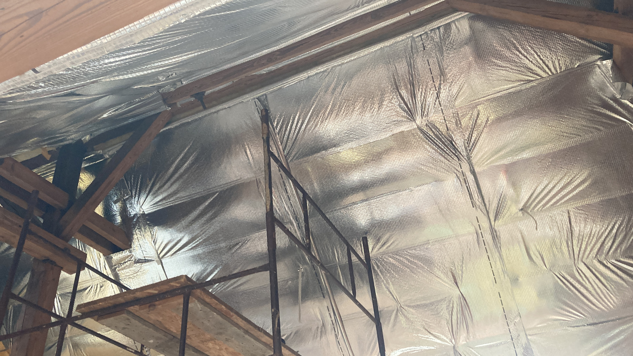 Srebrna folia izolacyjna zamontowana na drewnianej konstrukcji dachu poddasza, widoczny fragment rusztowania. Odbijające światło wykończenie.