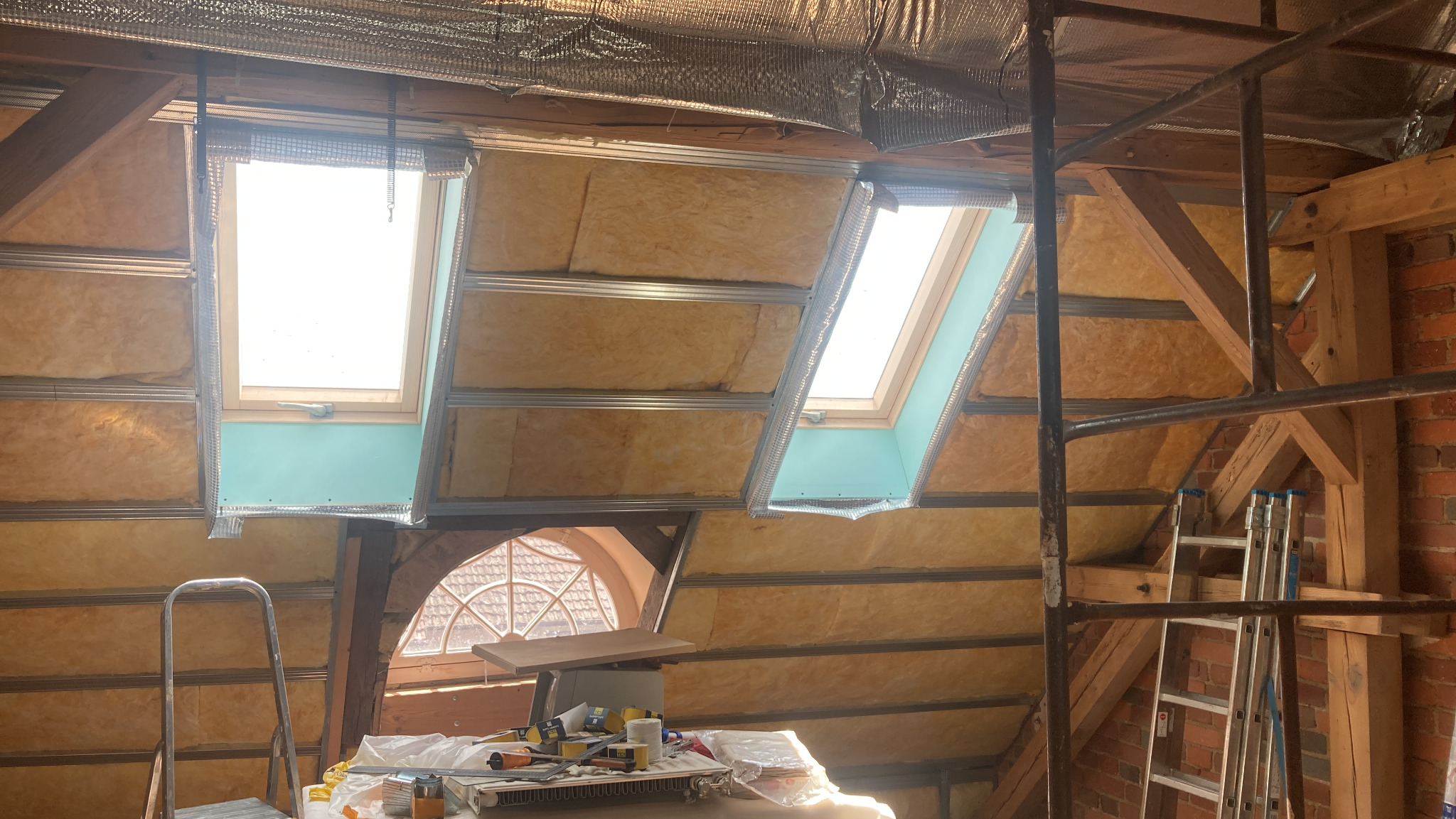 Poddasze z oknami dachowymi w trakcie izolacji wełną mineralną, widoczne profile i folia paroizolacyjna, rusztowanie i narzędzia pracy.