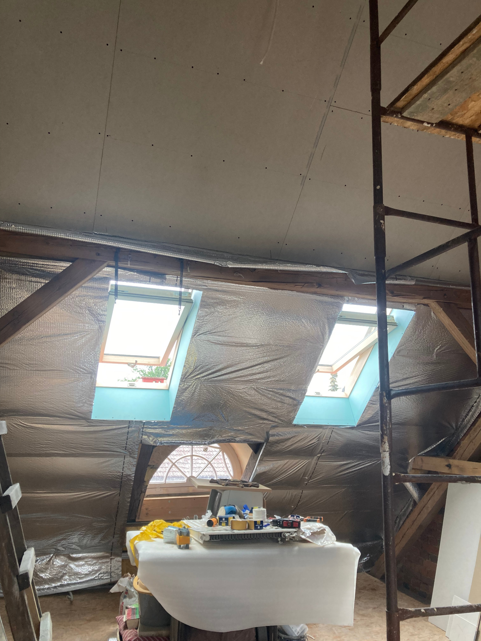 Poddasze w trakcie izolacji: folia aluminiowa na skośnych ścianach, okna dachowe, płyty karton-gips na suficie, rusztowanie. Widoczne narzędzia i materiały budowlane.