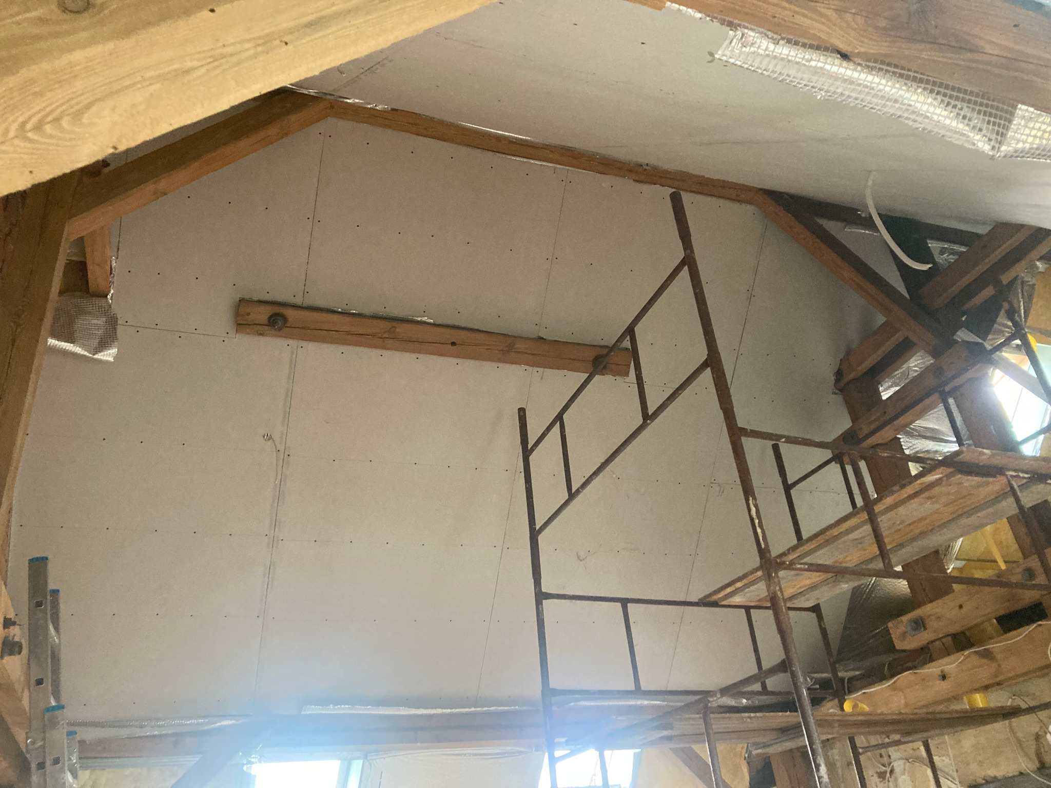 Skośny sufit poddasza z płyt kartonowo-gipsowych, widoczne drewniane belki konstrukcyjne i rusztowanie. Praca w toku, widok z dołu.