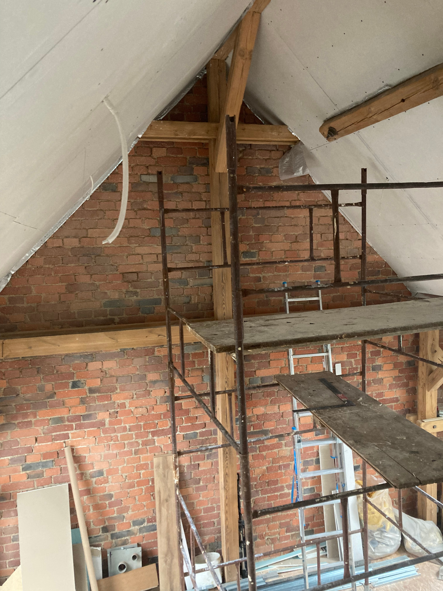 Skośny strych z odsłoniętą cegłą i płytami gipsowymi na suficie, widoczne rusztowanie i materiały budowlane, etap prac wykończeniowych.