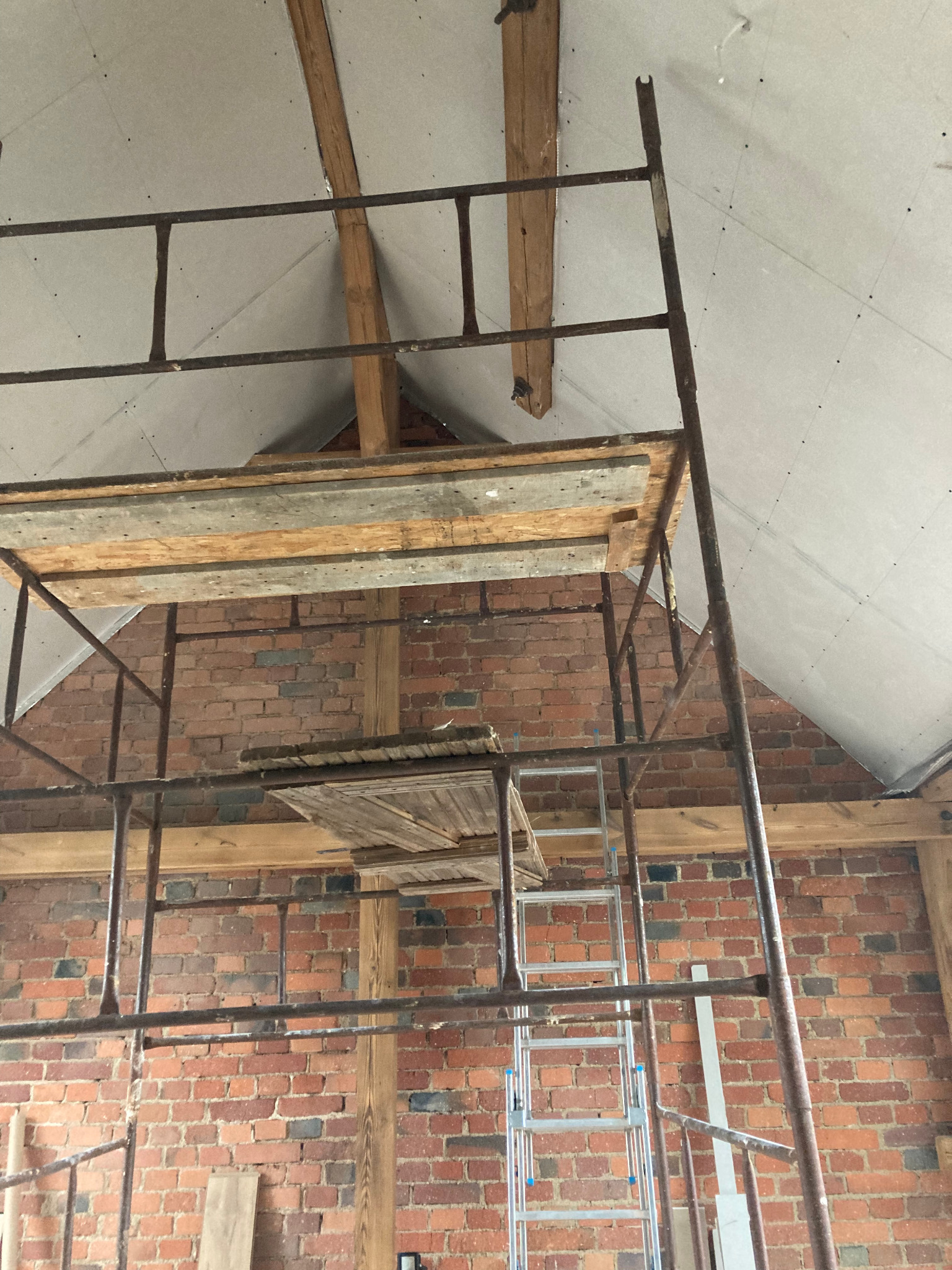 Montaż płyt karton gips na poddaszu: widok na rusztowanie, sufit z płyt, ceglaną ścianę i drewniane elementy konstrukcyjne. Praca w toku, surowe wykończenie.