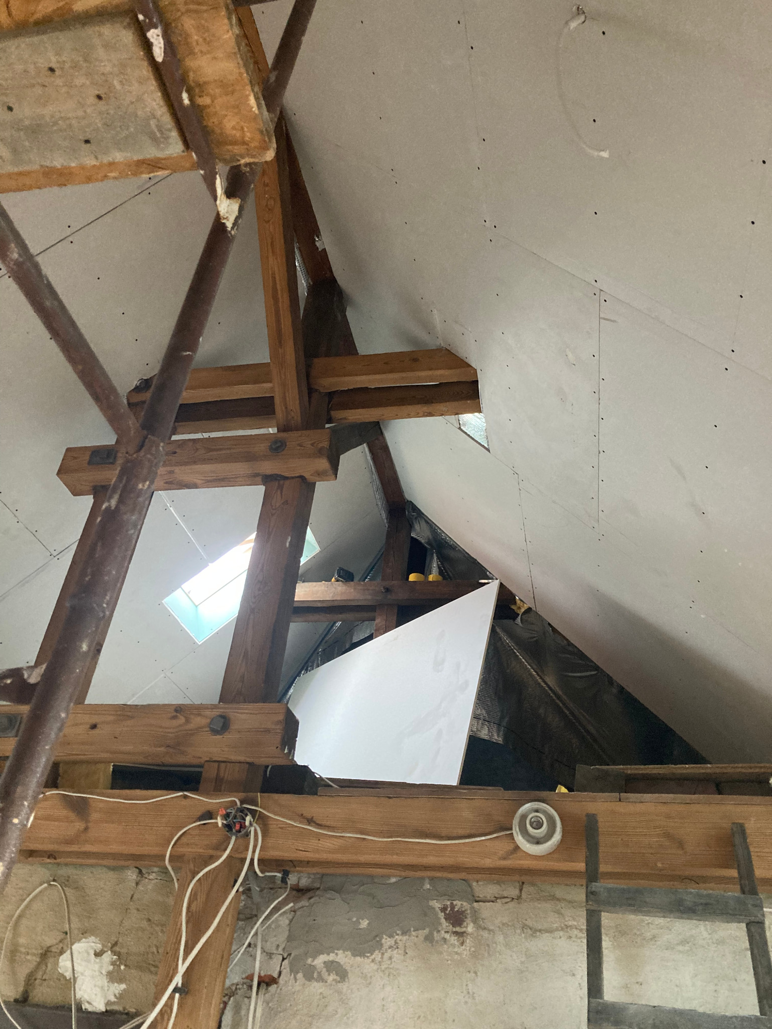 Poddasze w trakcie wykańczania płytami karton-gips, widoczne drewniane elementy konstrukcyjne, rusztowanie i instalacja elektryczna. Ujęcie z dołu na skośny sufit.