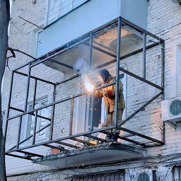 Prszem&Slav - Spawanie metalowej konstrukcji balkonu przez spawacza w masce ochronnej, na tle białej ceglanej ściany budynku. Widoczne iskry i dym podczas pracy.