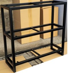 Prszem&Slav - Czarna, metalowa konstrukcja regału z trzema poziomami, spawana z profili, na tle kartonowych pudeł i betonowej posadzki.