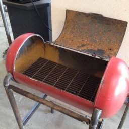 Prszem&Slav - Spawany grill z czerwonej butli gazowej, z rusztem w środku i otwartą pokrywą, na metalowym stelażu. Widoczne ślady rdzy i spawania.