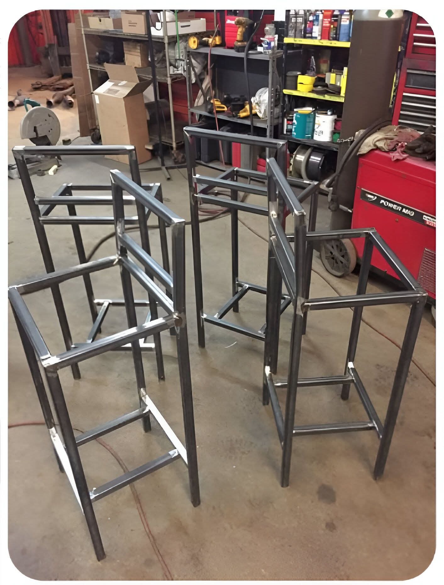 Metalowe ramy krzeseł barowych w warsztacie, spawane łączenia widoczne, czerwona spawarka w tle, surowy industrialny wygląd, przygotowane do dalszej obróbki.