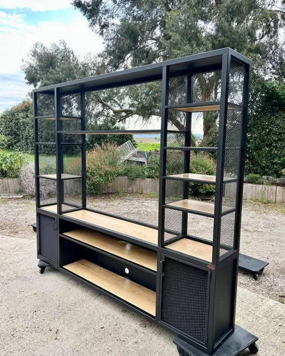 Czarna, metalowa konstrukcja regału z drewnianymi półkami, na kółkach, z ażurowymi ściankami, na zewnątrz, z widocznym tłem zieleni i fragmentem nieba.