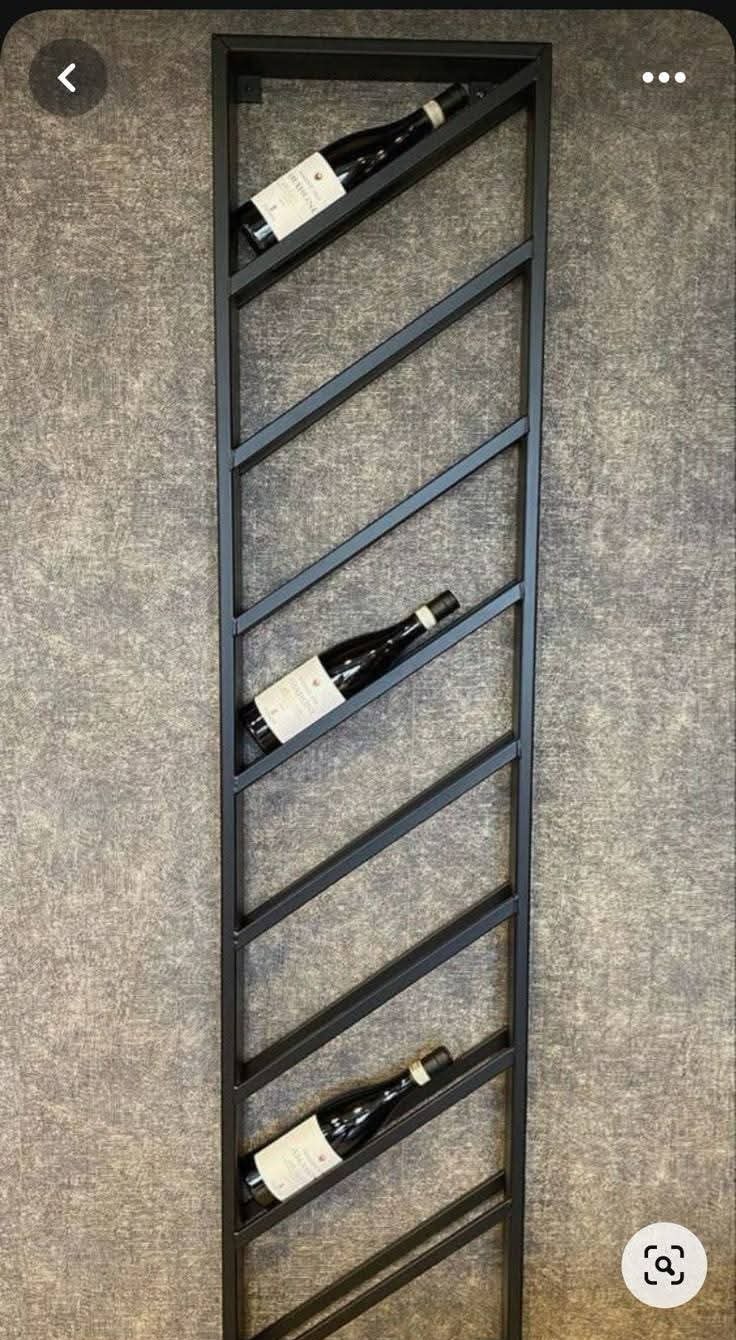 Metalowy, czarny regał na wino, przymocowany do ściany. Ukośne półki mieszczą trzy butelki wina z etykietami. Minimalistyczny design.