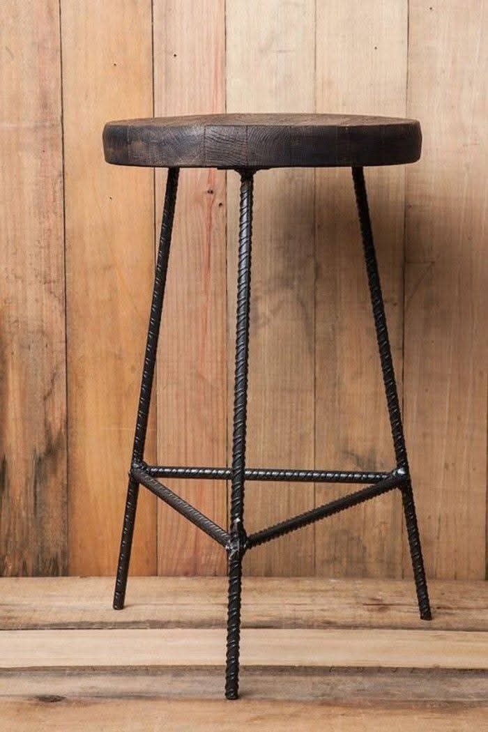 Stołek barowy z drewnianym siedziskiem i metalową konstrukcją z prętów żebrowanych, spawany, na tle drewnianej ściany. Surowy, industrialny styl.