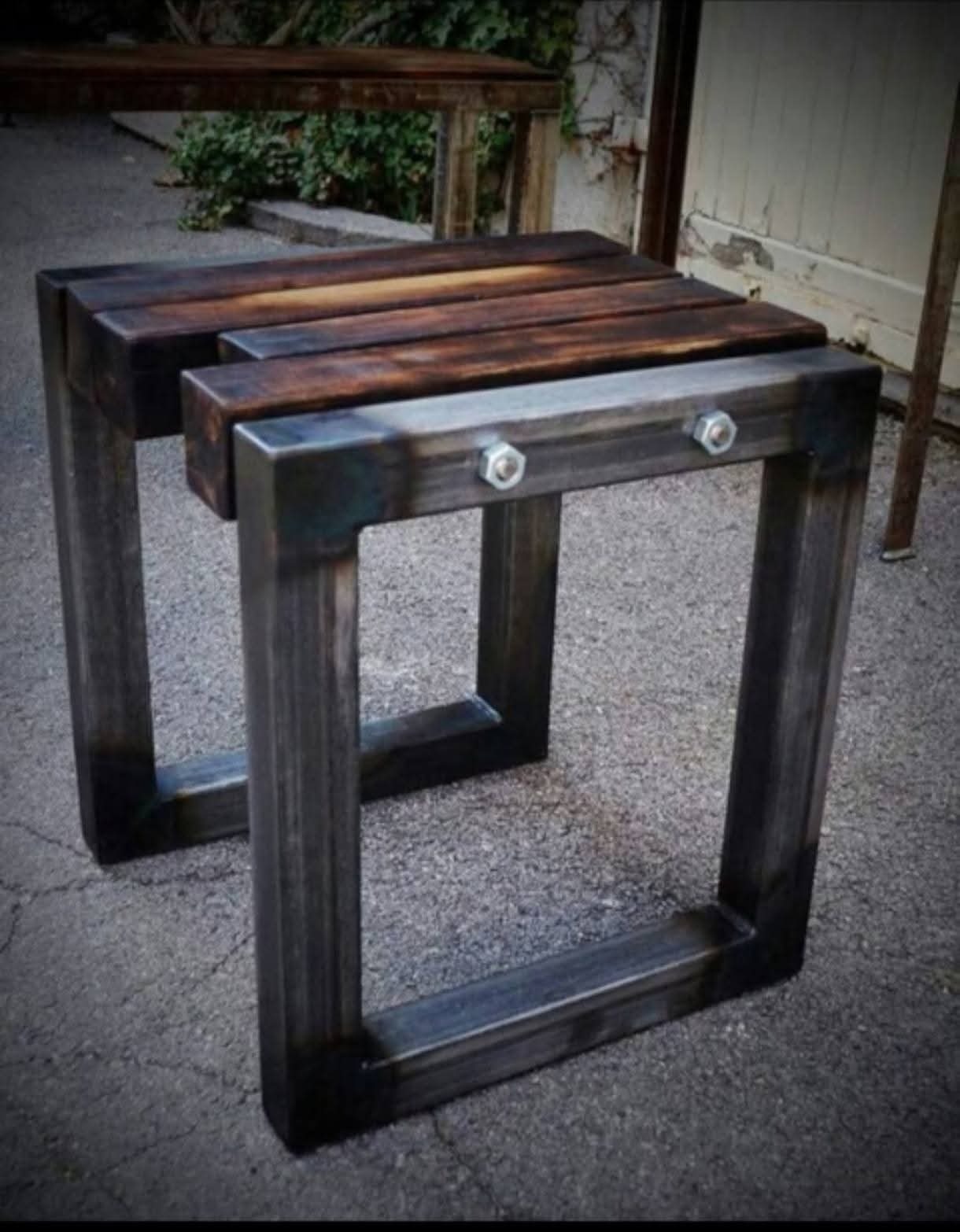Dwa industrialne stołki z ciemnego drewna i metalu, połączone śrubami, na tle betonowej nawierzchni i drzwi. Widoczne detale spawów i faktura drewna.
