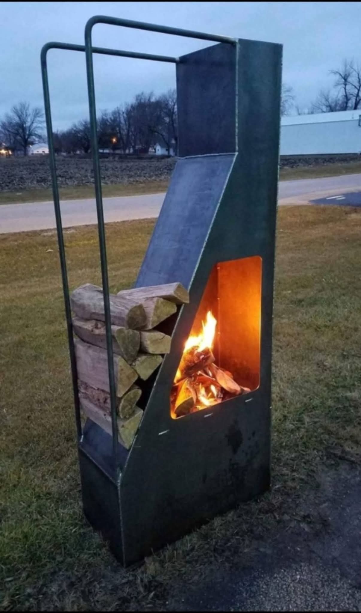 Metalowy kosz na drewno z paleniskiem na zewnątrz. W koszu ułożone drewno, w palenisku płonie ogień. Konstrukcja spawana, surowy metal, tło: trawa, droga.