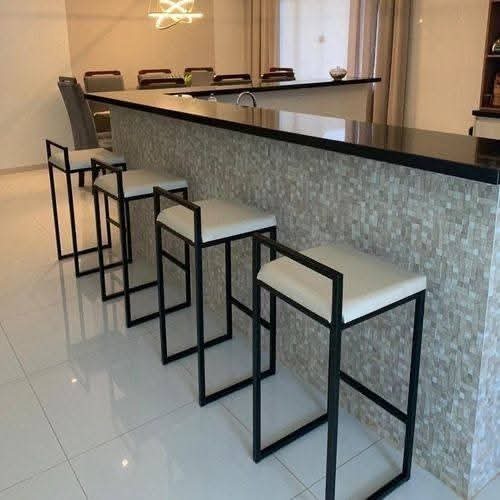 Rząd nowoczesnych, czarnych hokerów z białymi siedziskami ustawionych przy barze wykończonym mozaiką. Minimalistyczny design, jasne wnętrze, płytki podłogowe.