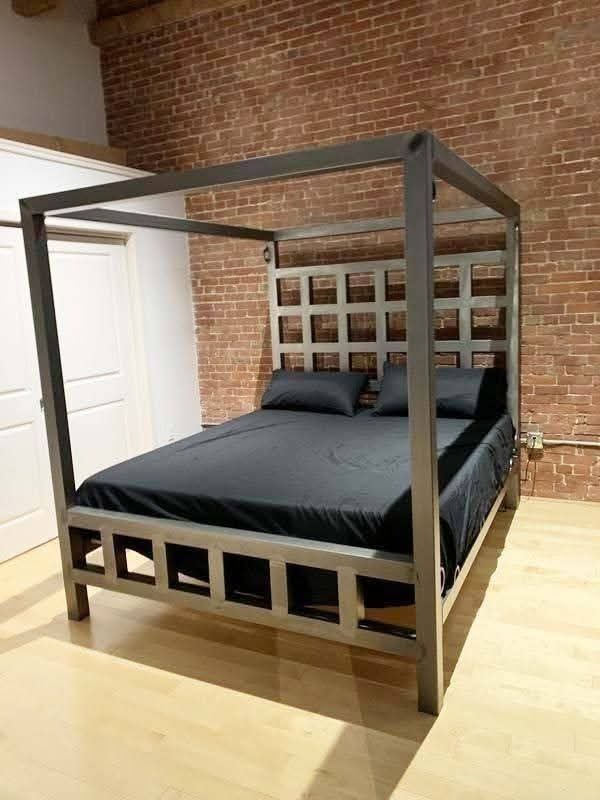 Metalowe łóżko z baldachimem o surowym, industrialnym designie, z kratownicowym wezgłowiem, na tle ceglanej ściany i jasnej podłogi.