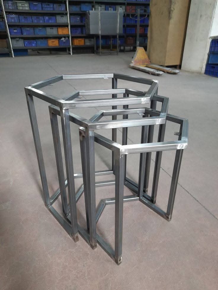 Surowe, metalowe konstrukcje w kształcie heksagonów, ustawione jedna w drugiej, tworzące zestaw stolików. Widoczne spawy i surowy metal, tło magazynowe.