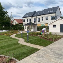 Grupa BNK BARTŁOMIEJ NOWAKOWSKI - Zadbany ogród z trawnikiem, ścieżką z kamienia i rabatami kwiatowymi. W tle biała altana, budynek w stanie surowym i osoba podlewająca rośliny. Całość w letniej scenerii.