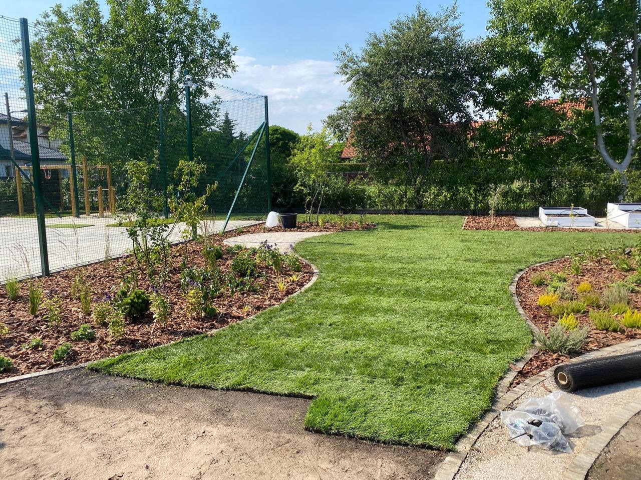 Układanie trawy z rolki na działce z rabatami i korą. Widoczne narzędzia i rolka trawy. Trawa świeża, soczysta zieleń. Dzień słoneczny, błękitne niebo.