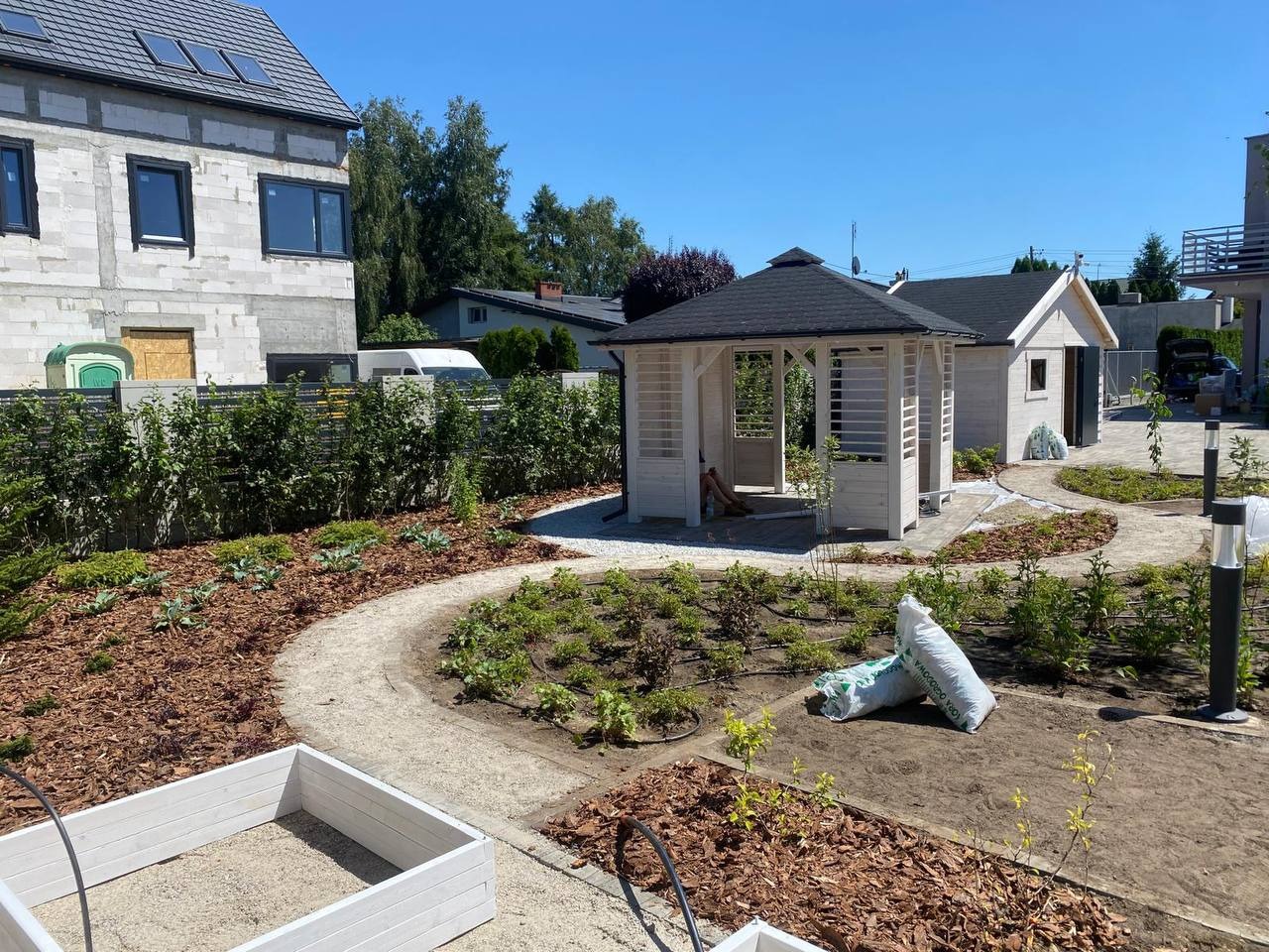 Nowo założony ogród z białą altaną, ścieżką z kamyków i rabatami z korą. Widoczne worki z ziemią i system nawadniania. Dom w tle w trakcie budowy.