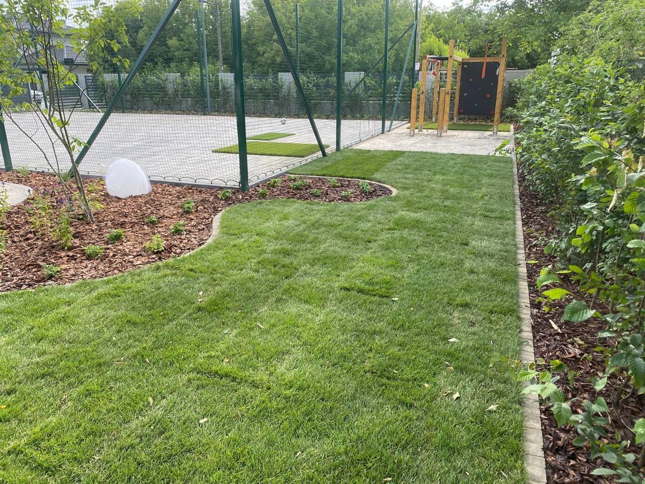 Świeżo założony trawnik z rolki, obrzeża z kostki, kora i nasadzenia w tle boiska sportowego i placu zabaw. Widok z góry, słoneczny dzień.
