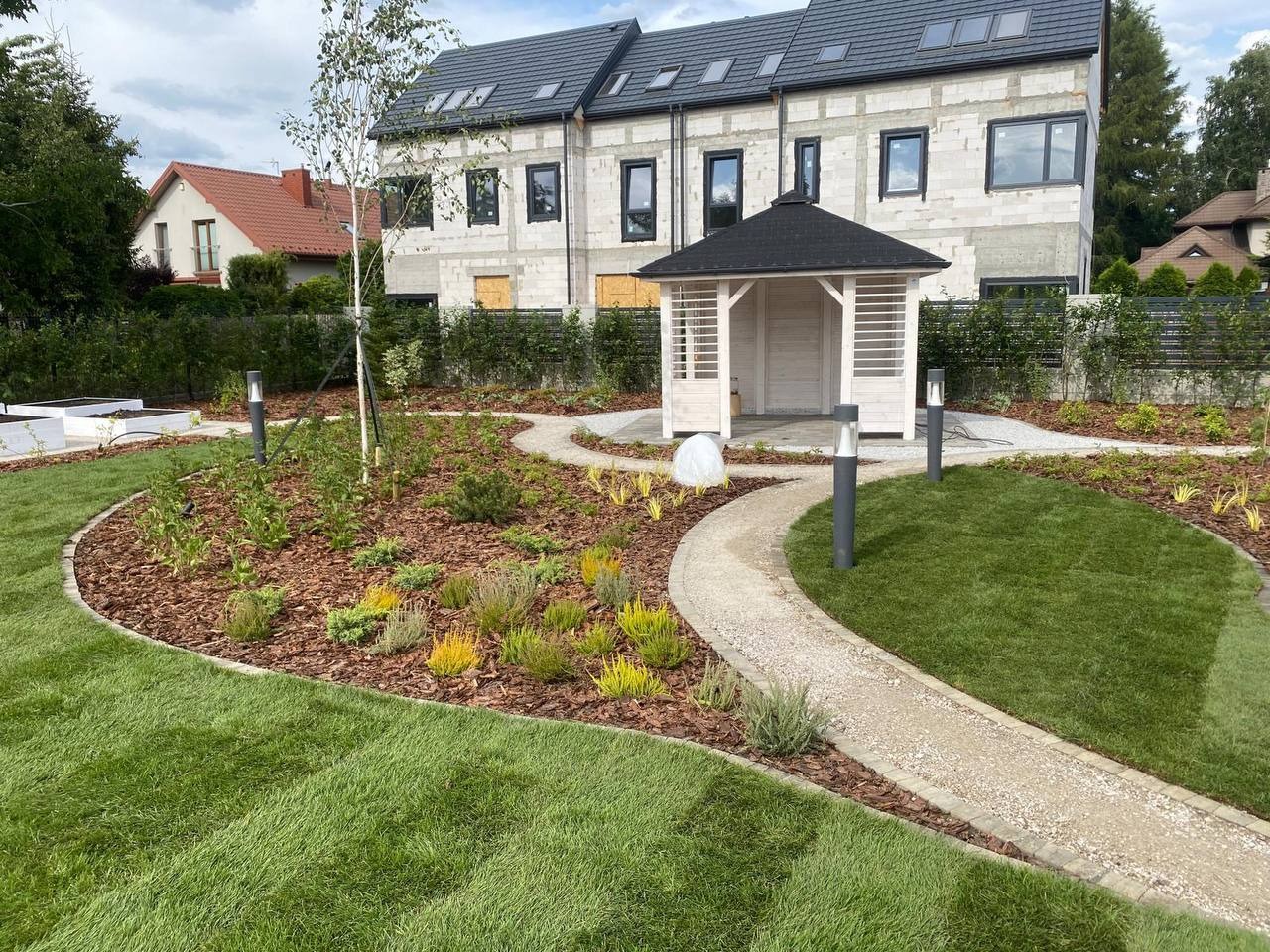 Nowoczesny ogród z altaną i ścieżką z kamienia, świeżo założony trawnik, rabaty z korą i niskimi roślinami, oświetlenie ogrodowe, budynek w tle.