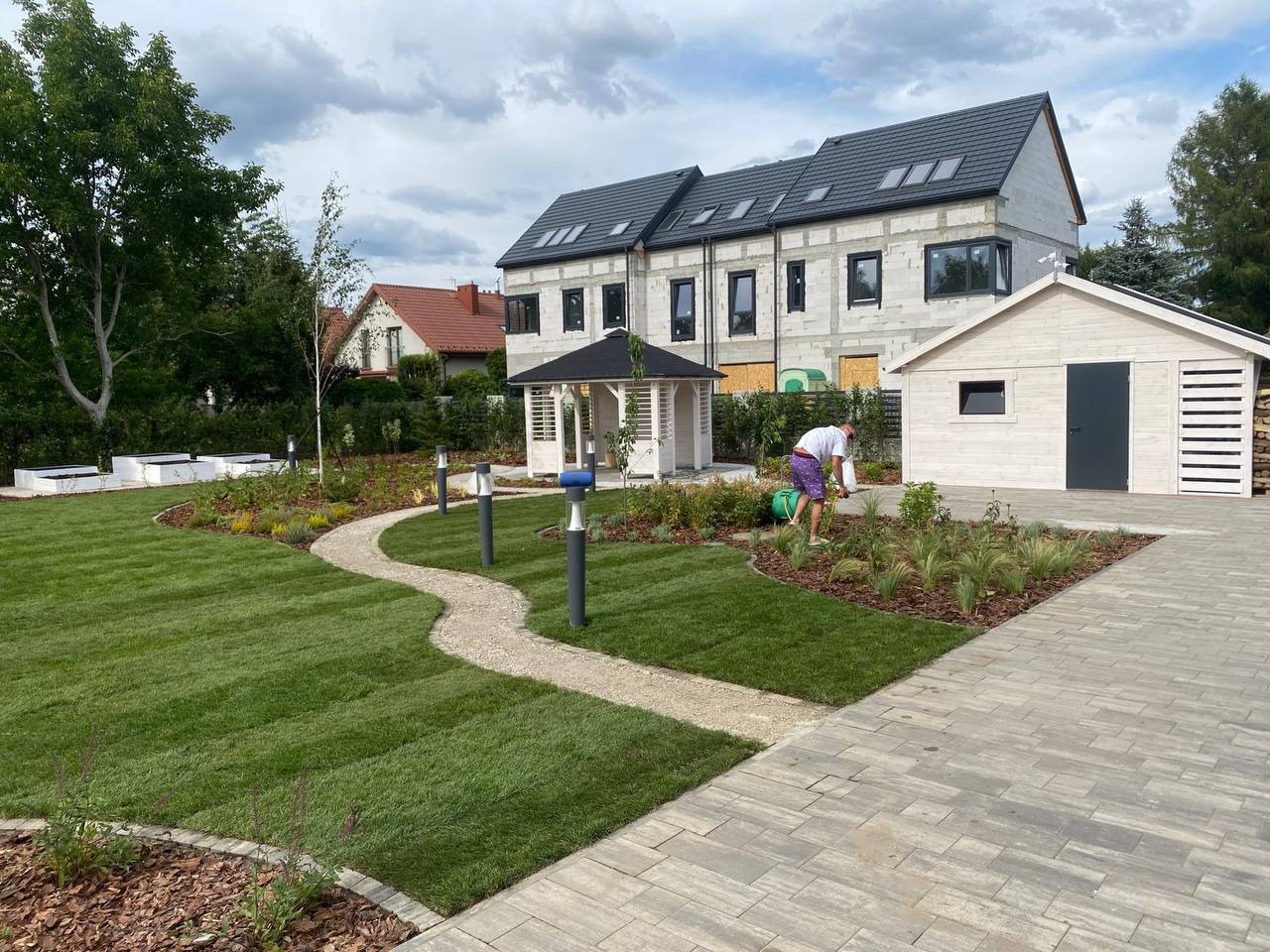 Zadbany ogród z trawnikiem, ścieżką z kamienia i rabatami kwiatowymi. W tle biała altana, budynek w stanie surowym i osoba podlewająca rośliny. Całość w letniej scenerii.