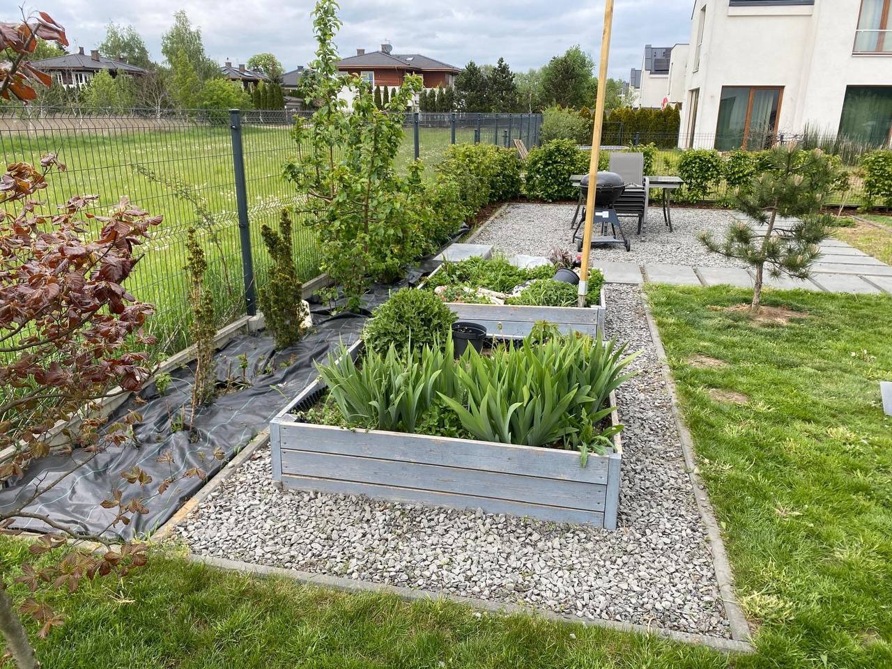 Ogród z drewnianymi rabatami, wysypany kamieniem, z młodymi roślinami i drzewami. W tle trawnik, ogrodzenie i dom. Widok na zagospodarowany teren.