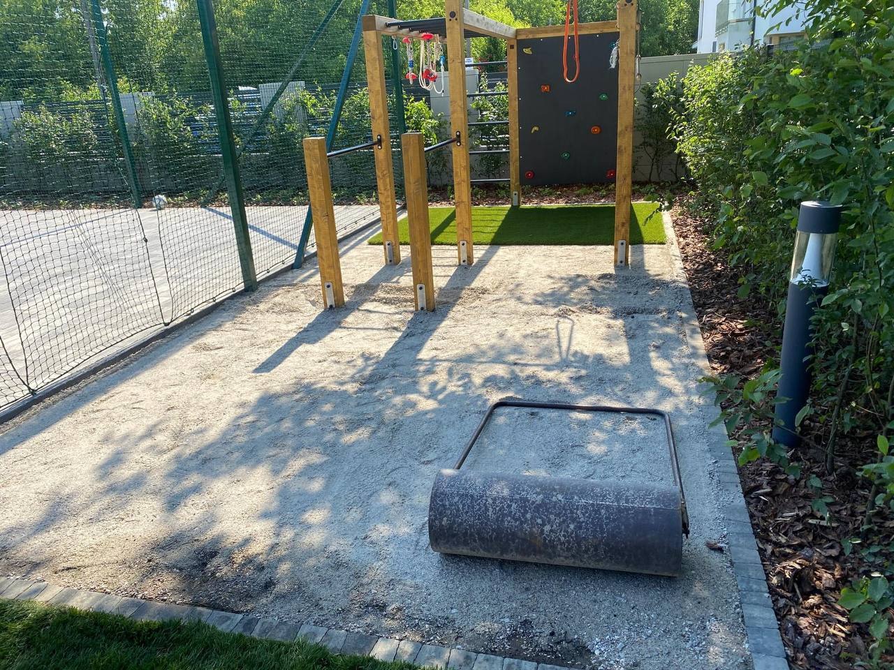 Przygotowanie terenu pod plac zabaw: ubita ziemia, walec, elementy konstrukcji, siatka ogrodzeniowa. Widok z góry, słoneczny dzień.