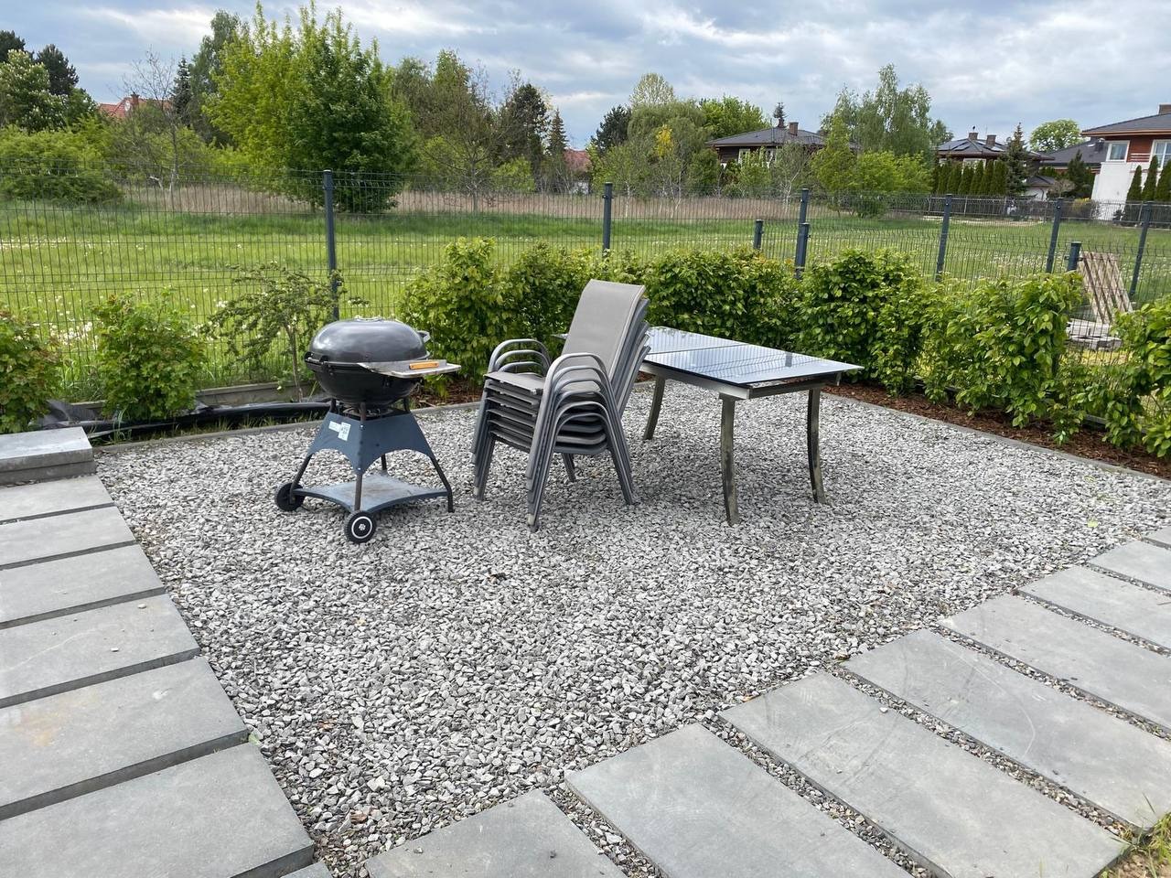 Ukończony taras z kamiennym podłożem, obrzeżami z płyt, grillem, stołem i krzesłami. Widok na zadbany ogród i dom w tle, pochmurne niebo.