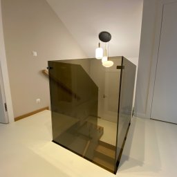 Kamil Wojciechowski - Szklana balustrada w nowoczesnym wnętrzu, widok na schody. Minimalistyczny design, neutralna kolorystyka ścian i podłogi, wisząca lampa nad balustradą.