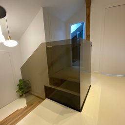 Kamil Wojciechowski - Szklana balustrada schodowa w nowoczesnym wnętrzu. Minimalistyczny design, neutralna kolorystyka, drewniane schody i białe drzwi. Widok z dołu, oświetlenie sufitowe.