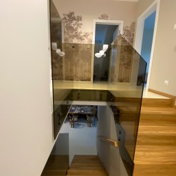 Kamil Wojciechowski - Nowoczesna, brązowa balustrada ze szkła na schodach z drewna, widok z góry. W tle tapeta z motywem drzew i widoczny fragment jadalni.