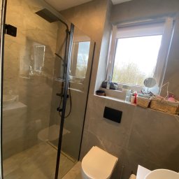 Kamil Wojciechowski - Nowoczesna kabina prysznicowa ze szklanymi drzwiami i czarnymi profilami w łazience wykończonej płytkami imitującymi marmur. Widok na okno z roślinnością.