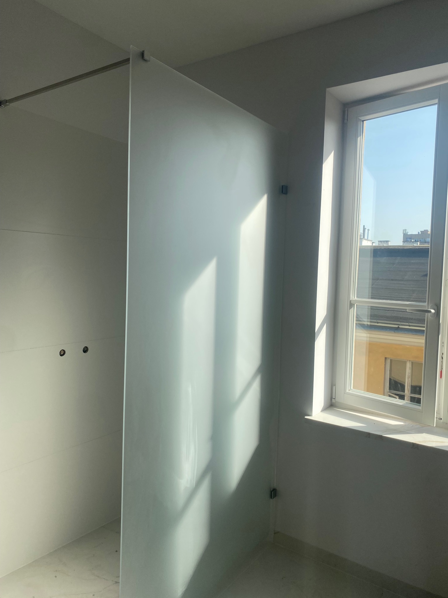 Minimalistyczna kabina prysznicowa ze szkła matowego w jasnej łazience z oknem. Widoczne mocowania i fragment instalacji elektrycznej.