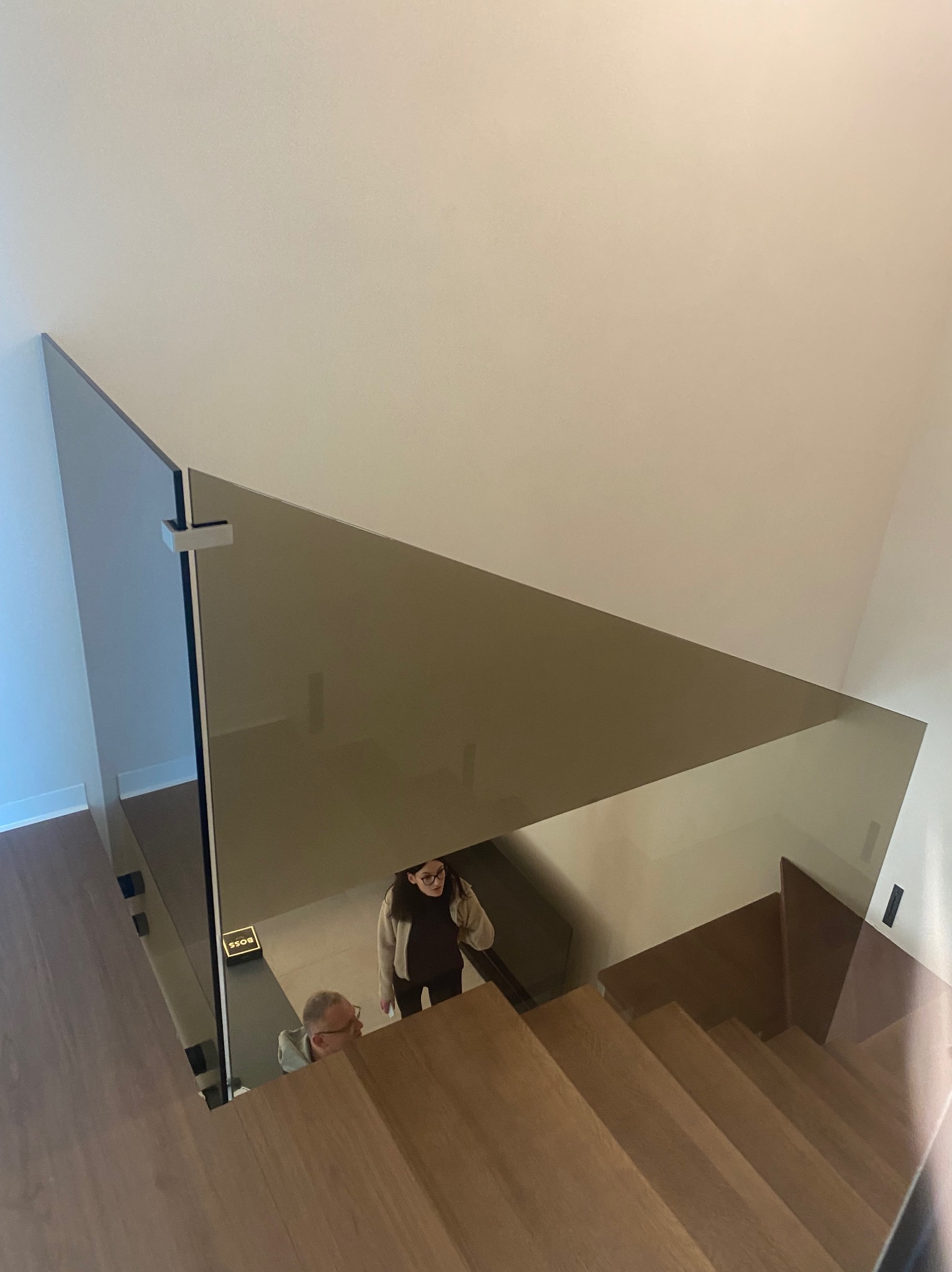 Szklana balustrada schodowa z brązowionego szkła, zamontowana w nowoczesnym domu. Widoczne drewniane schody i dwie osoby na górze. Minimalistyczny design.