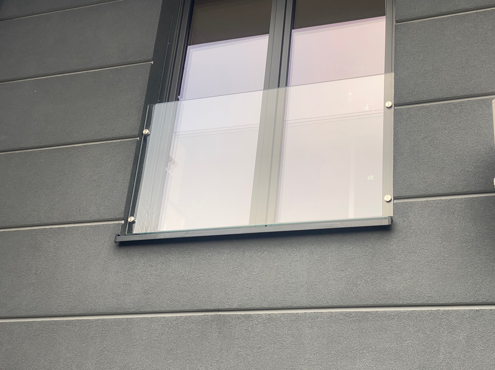 Szklana balustrada okienna zamontowana na szarej elewacji budynku, widoczna z bliska, mocowana na metalowych uchwytach.