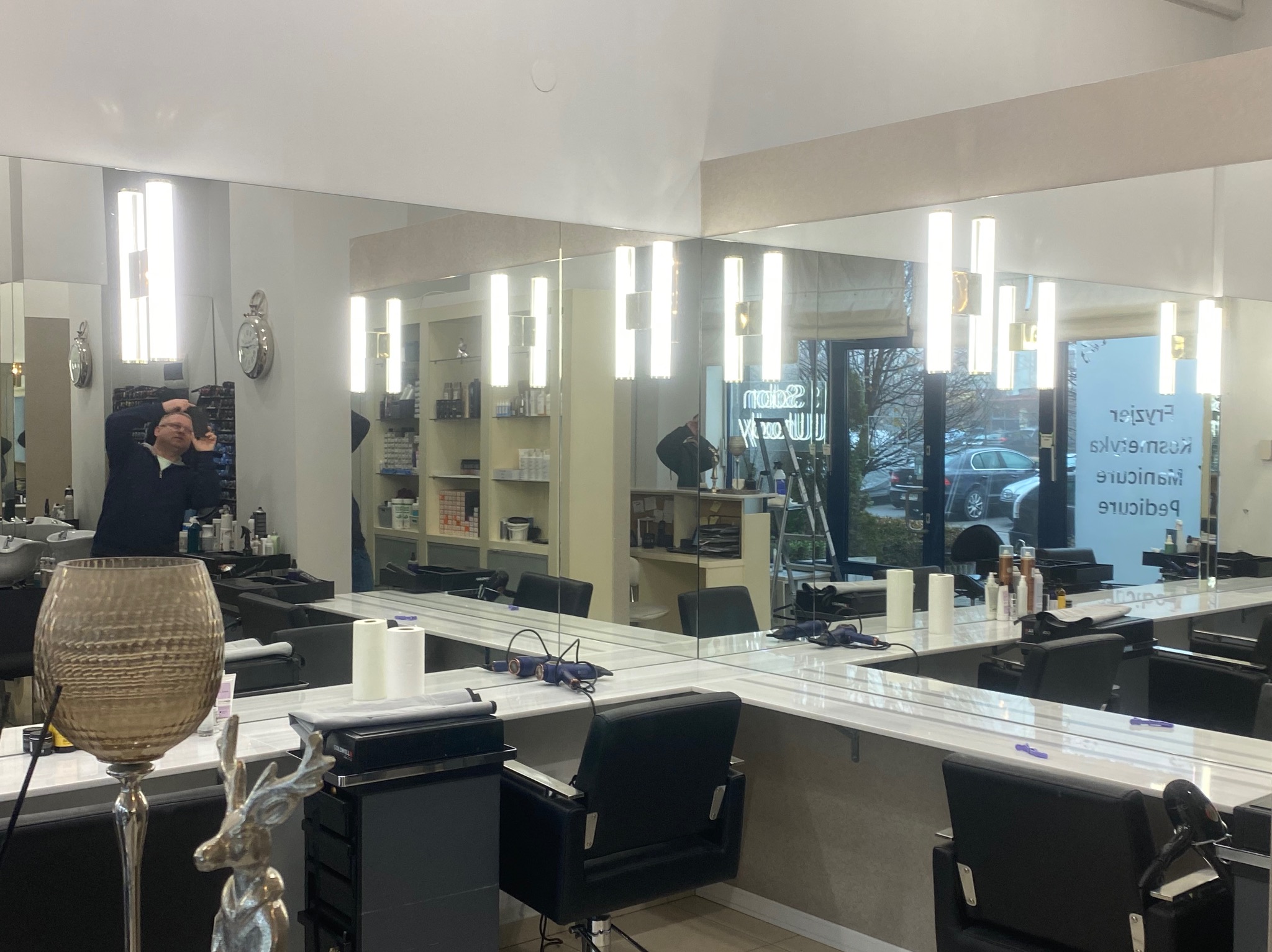 Nowoczesne lustra w salonie fryzjerskim z oświetleniem LED i stanowiskami pracy. W tle widoczny pracownik i wyposażenie. Dekoracyjny wazon na pierwszym planie.