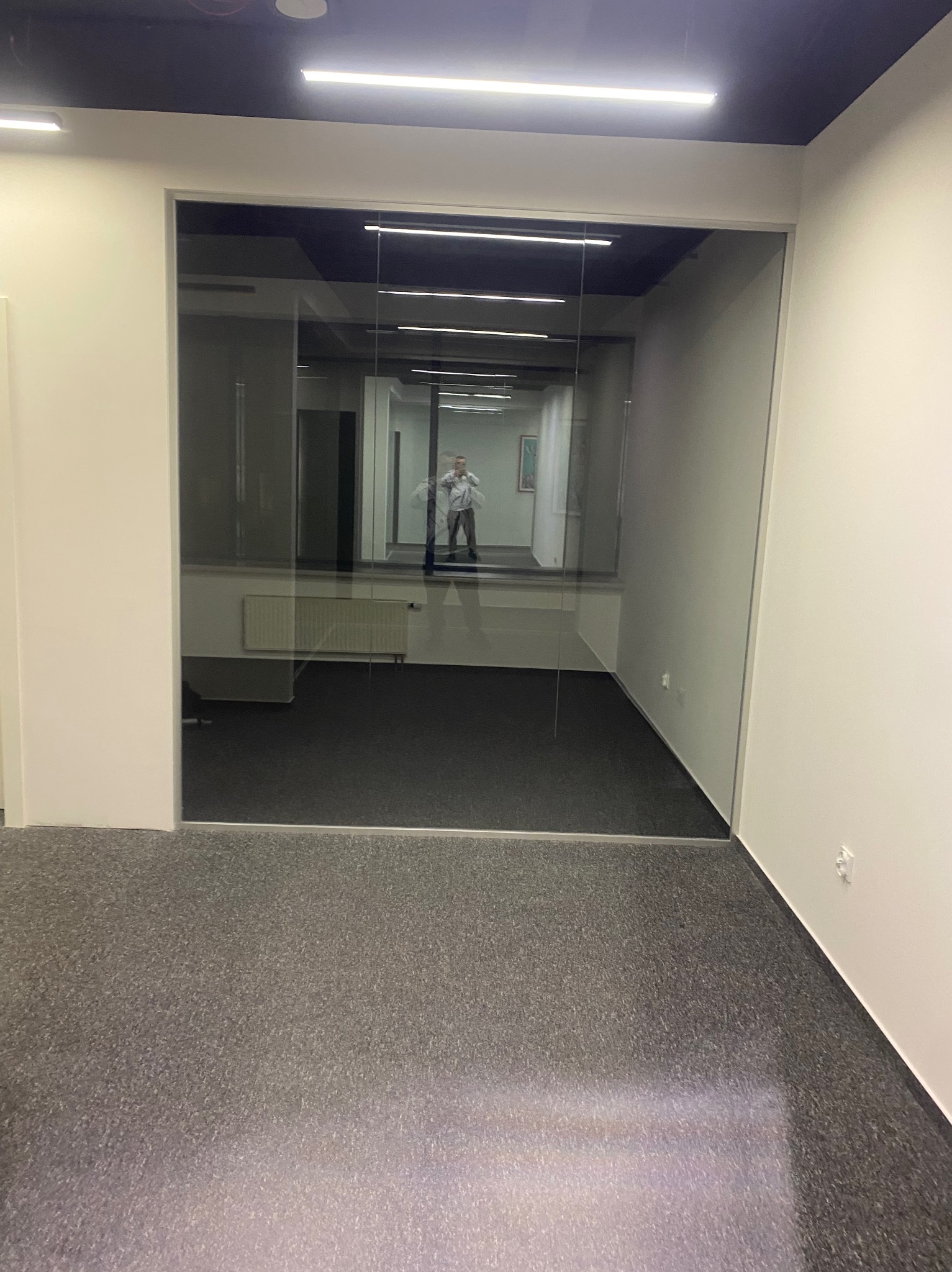 Szklane drzwi w biurze z widocznym odbiciem osoby. Minimalistyczny design, szara podłoga, białe ściany i sufit z oświetleniem LED. Nowoczesne wykończenie wnętrza.