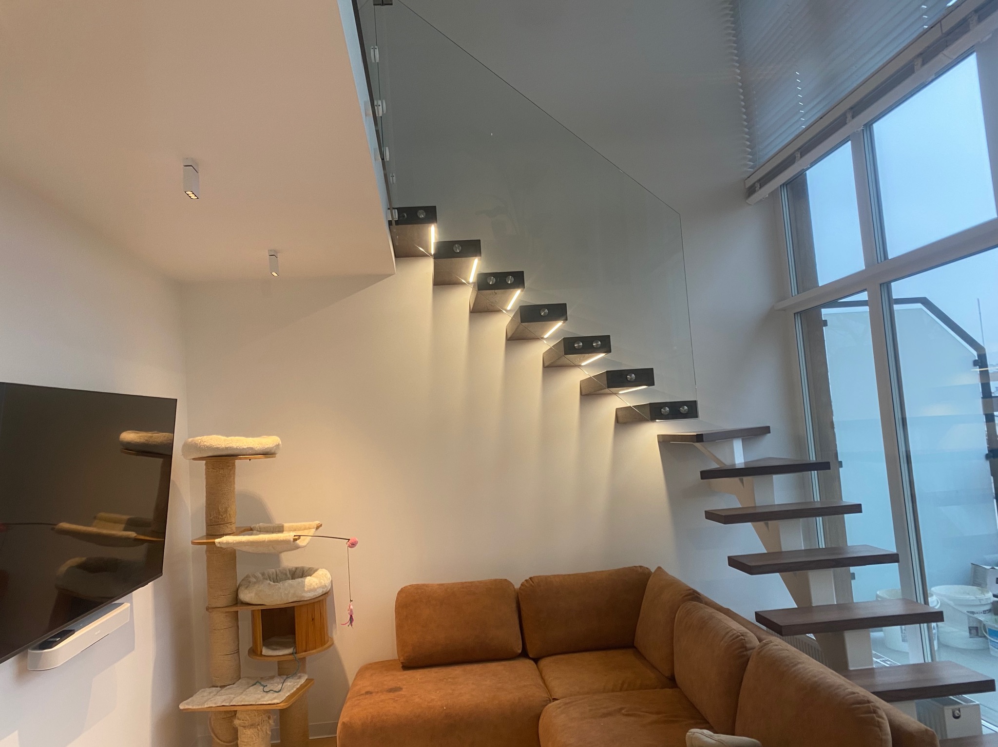 Nowoczesne schody wspornikowe z balustradą szklaną, oświetleniem LED i drewnianymi stopniami, widoczne z perspektywy salonu z brązową sofą i drapakiem dla kota.