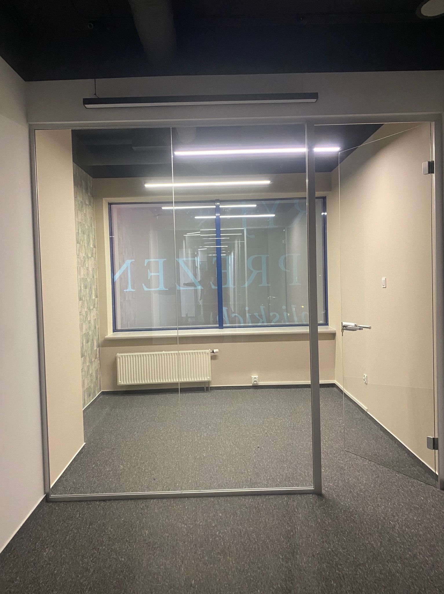 Szklane drzwi biurowe z widokiem na wnętrze. Minimalistyczny design, szara wykładzina, jasne ściany. Widoczne detale: klamka, zawiasy. Oświetlenie sufitowe.