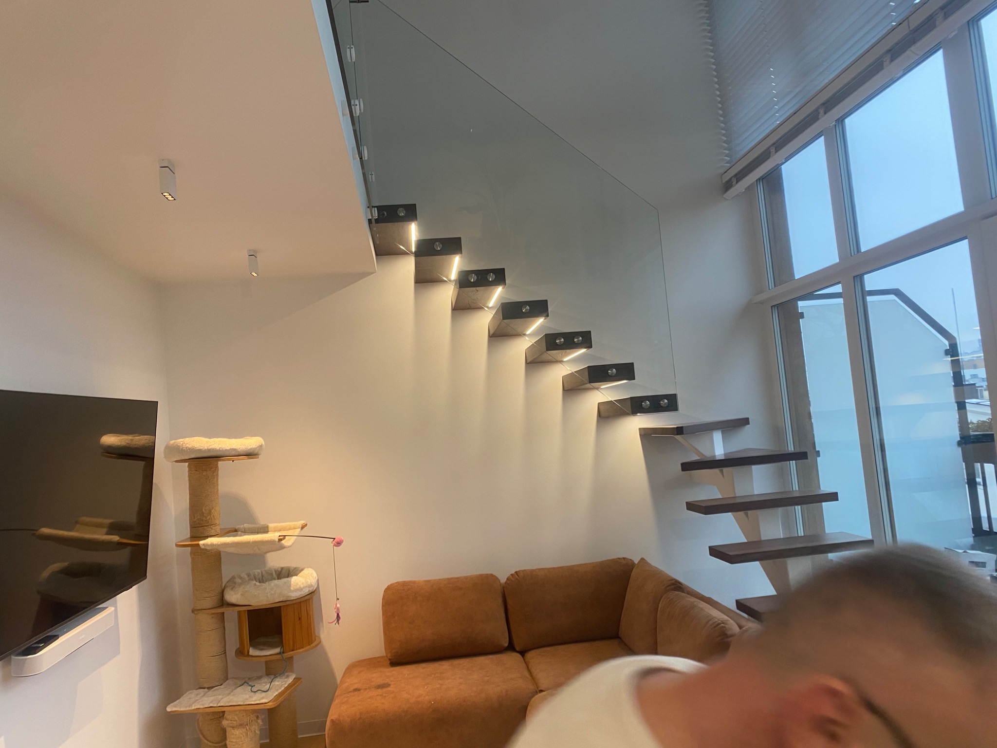 Nowoczesne schody z podświetlanymi stopniami i szklaną balustradą w jasnym wnętrzu. Widoczny fragment sofy i drapak dla kota.