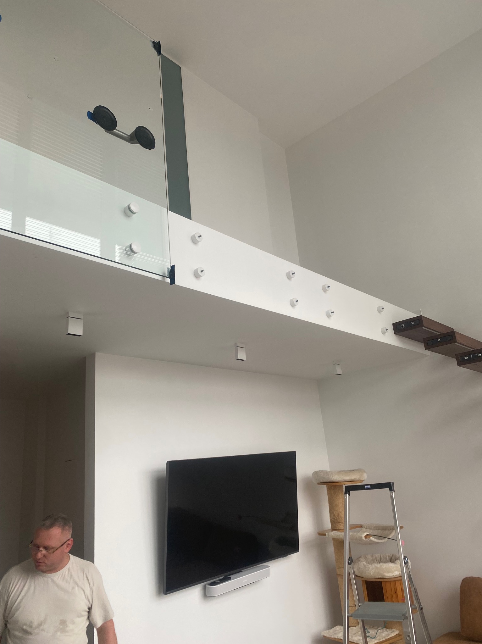 Szklana balustrada na antresoli, mocowana punktowo do białej belki. Widoczne przyssawki montażowe na szkle. W tle drabina i meble.