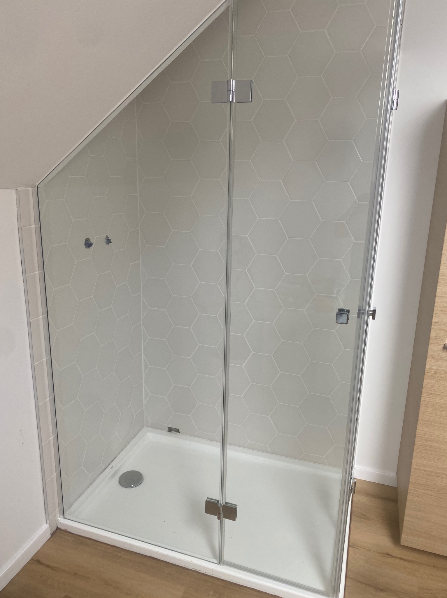 Szklana kabina prysznicowa z heksagonalnymi płytkami w łazience na poddaszu. Kabina z chromowanymi zawiasami i uchwytem, biały brodzik, drewnopodobna podłoga.