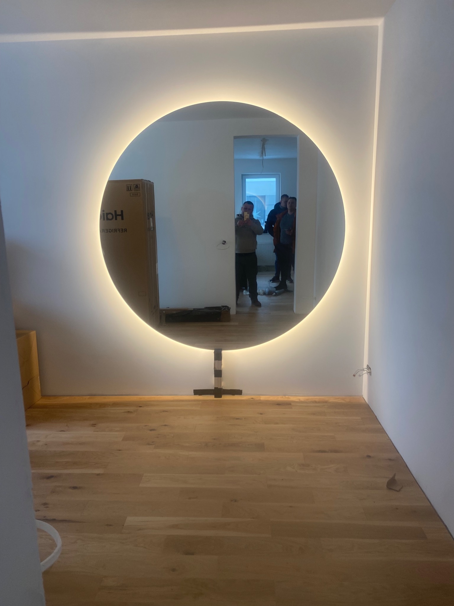 Okrągłe lustro z podświetleniem LED na metalowej podstawie, odbijające grupę osób w jasnym wnętrzu z drewnianą podłogą i kartonem Haier w tle.