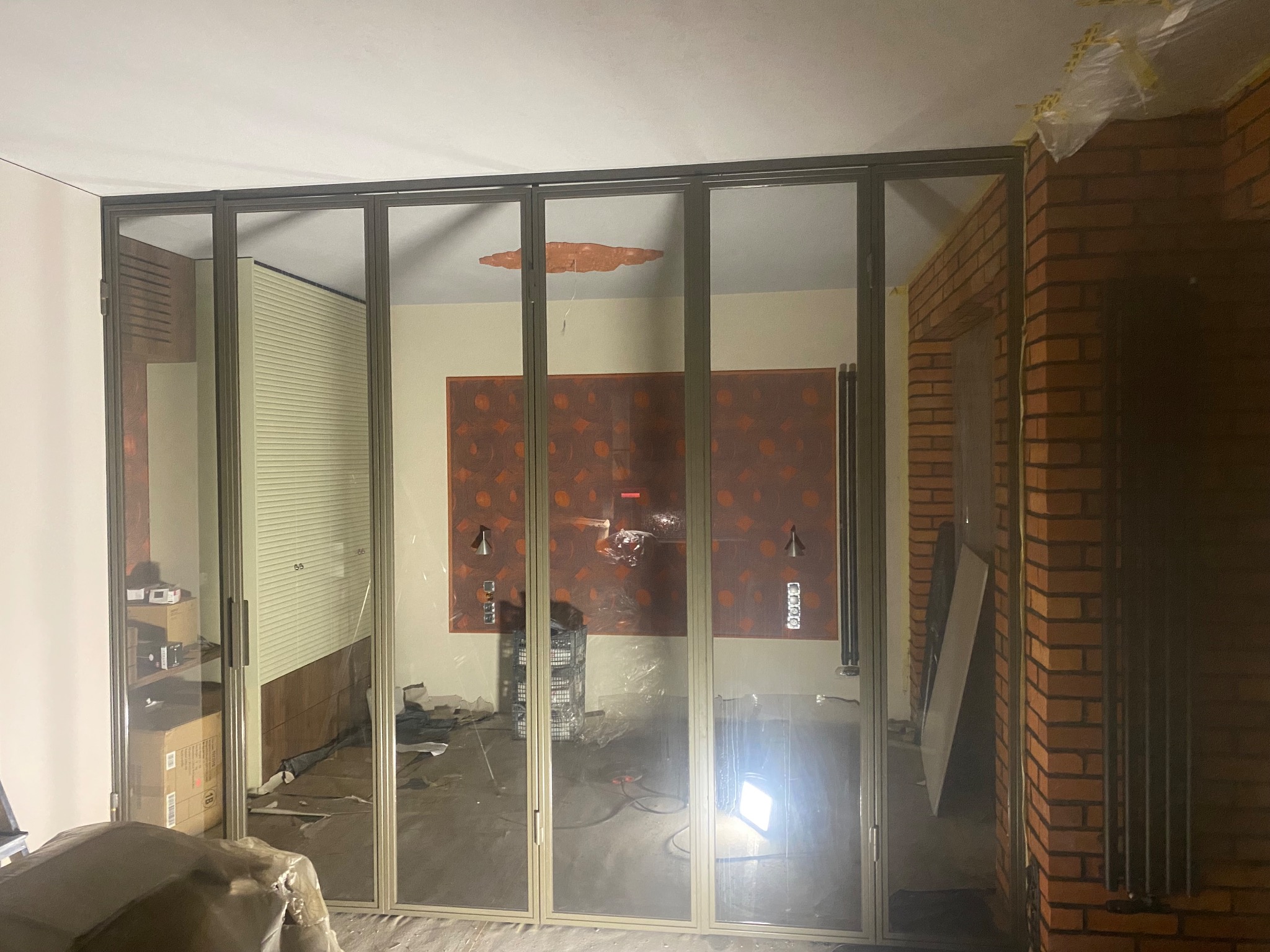 Szklane drzwi przesuwne z aluminiową ramą oddzielają pomieszczenia. Widoczny fragment ceglanej ściany i przygotowania do prac wykończeniowych.