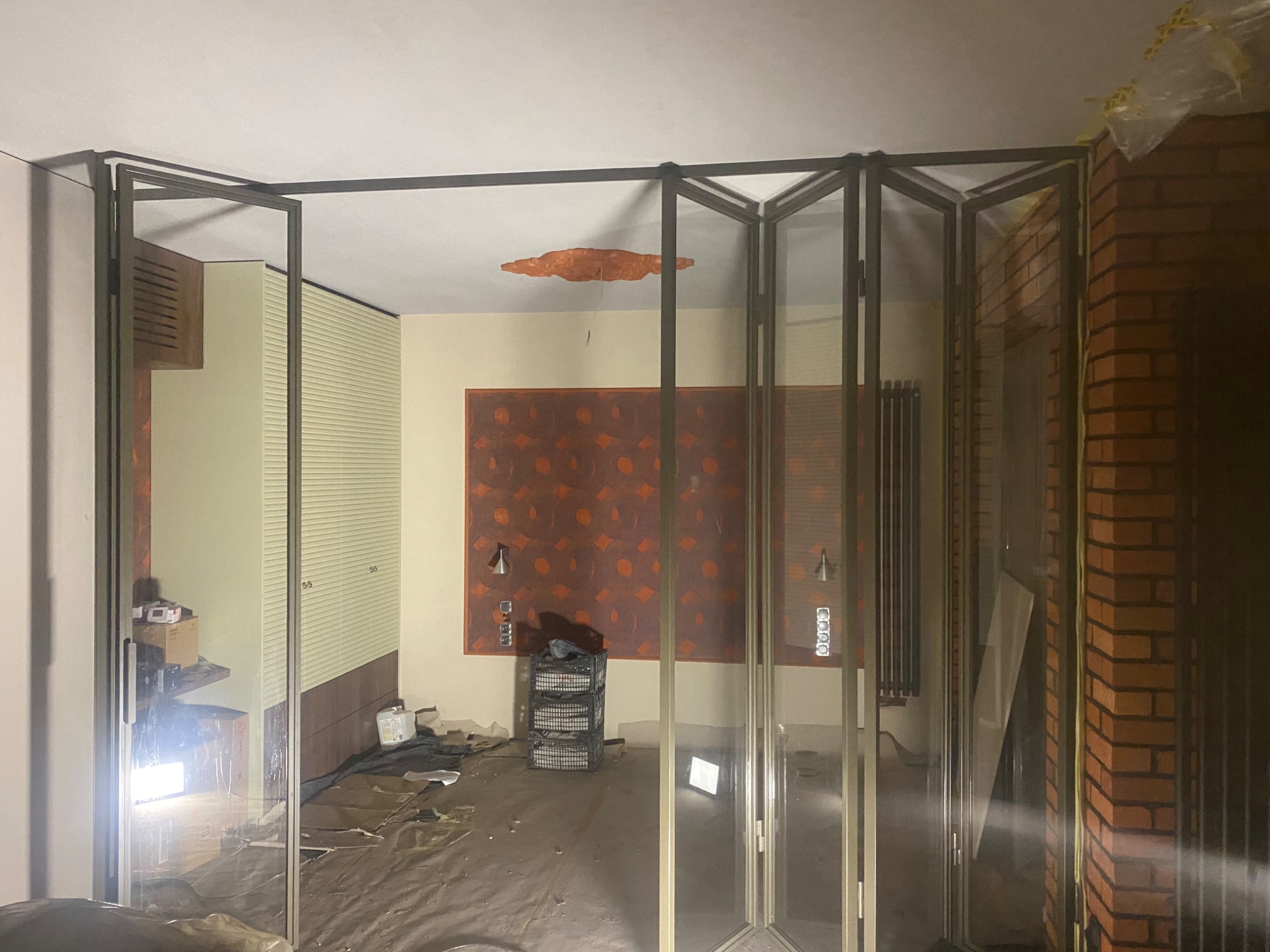 Składane drzwi szklane z ciemną ramą w trakcie montażu, widoczne zabezpieczenia na ścianie z cegły i podłodze. W tle ściana z abstrakcyjnym wzorem.