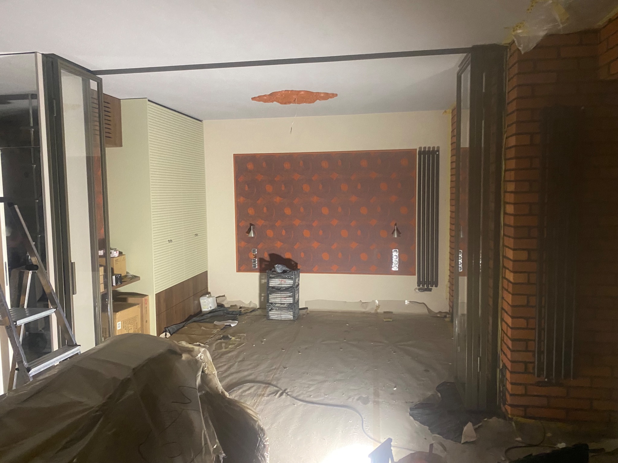 Wnętrze w trakcie remontu z rozsuwanymi drzwiami, zabezpieczoną podłogą i ceglanym wykończeniem ściany. Widoczne drabina i elementy wyposażenia.