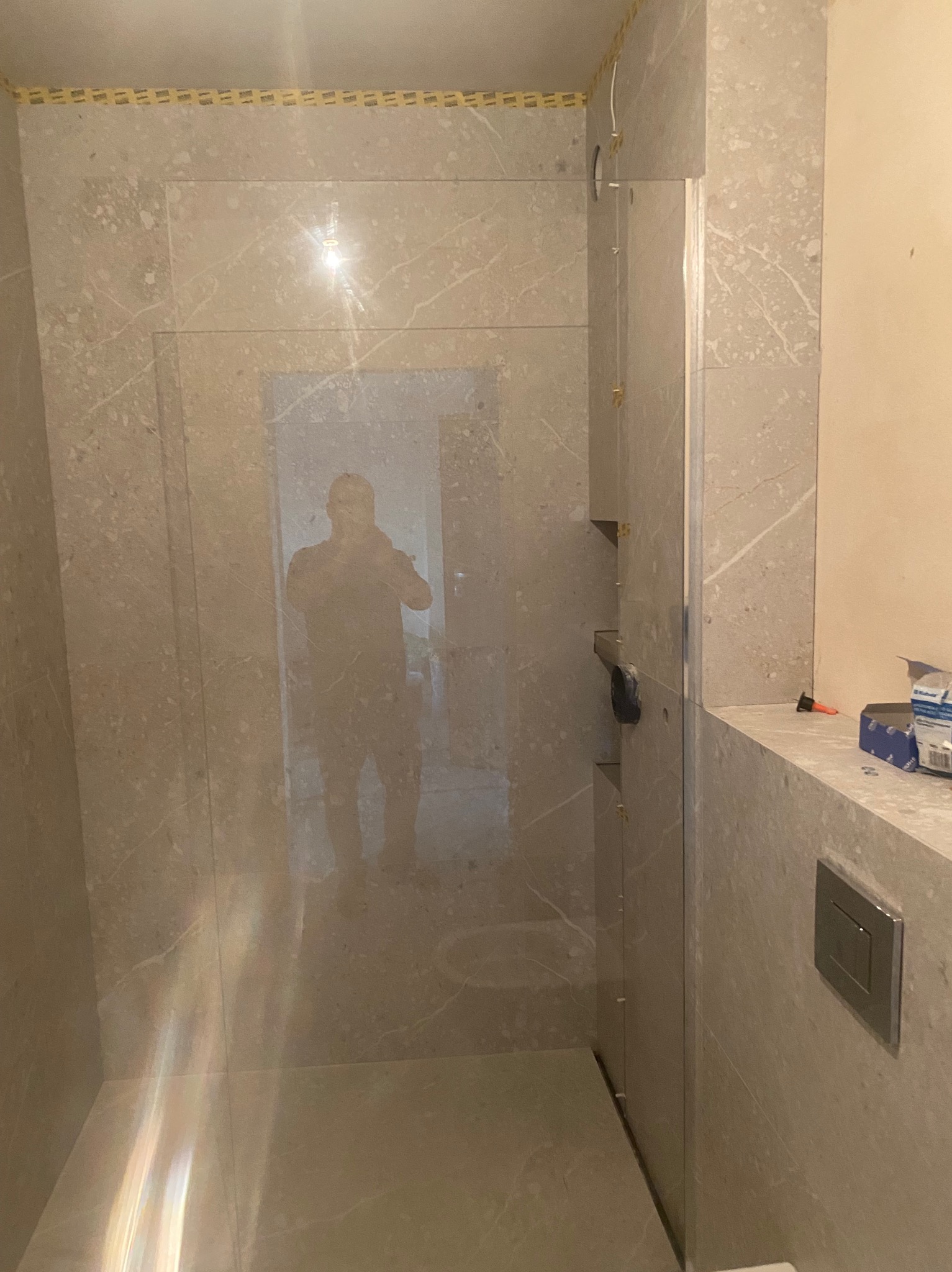 Szklana kabina prysznicowa z widocznym odbiciem osoby, wykończona płytkami imitującymi marmur. Widoczne elementy montażowe i akcesoria łazienkowe.