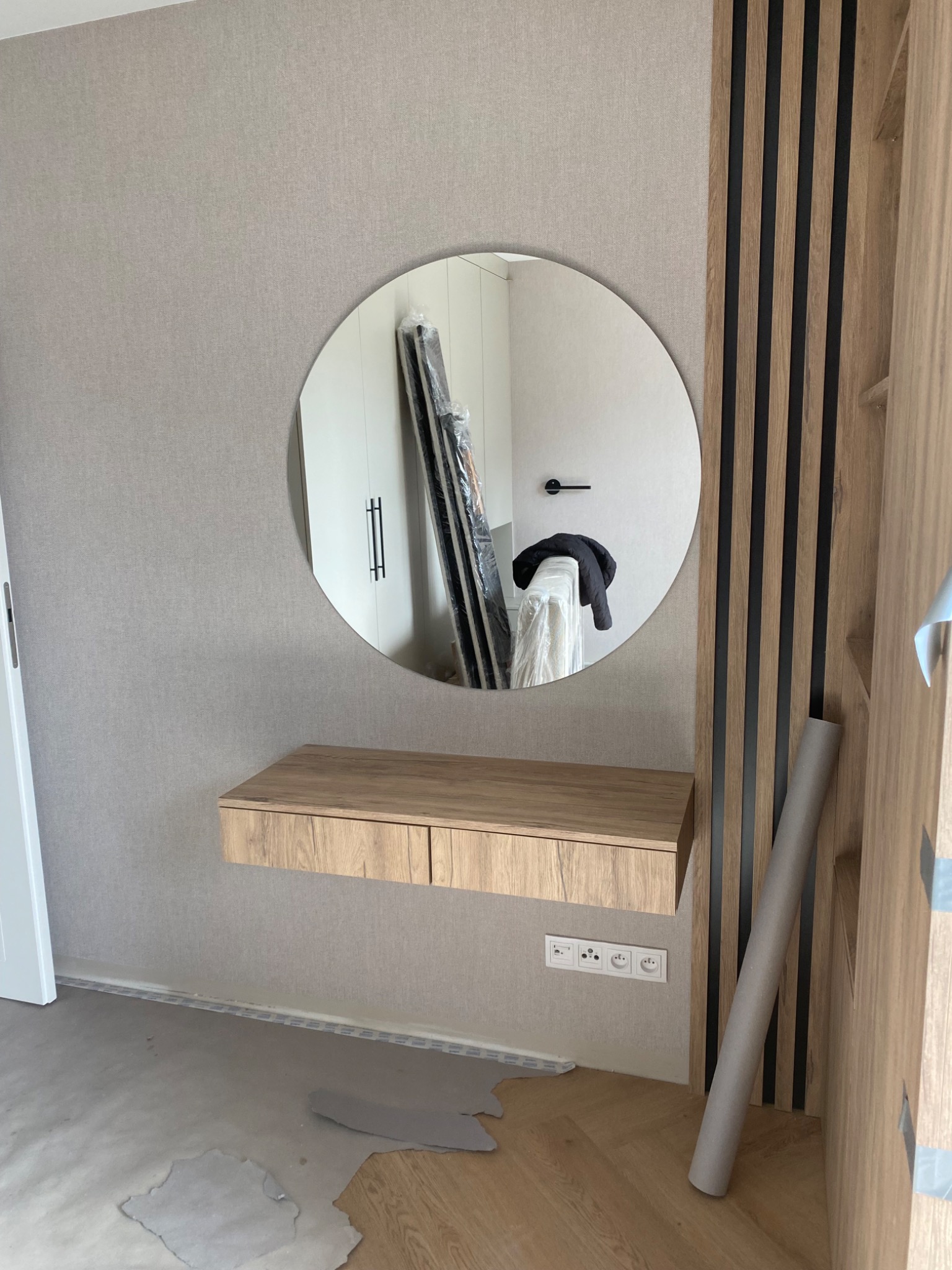 Owalne lustro z drewnianą konsolą w jasnym wnętrzu. Widoczne listwy dekoracyjne na ścianie i fragment podłogi w trakcie prac wykończeniowych.