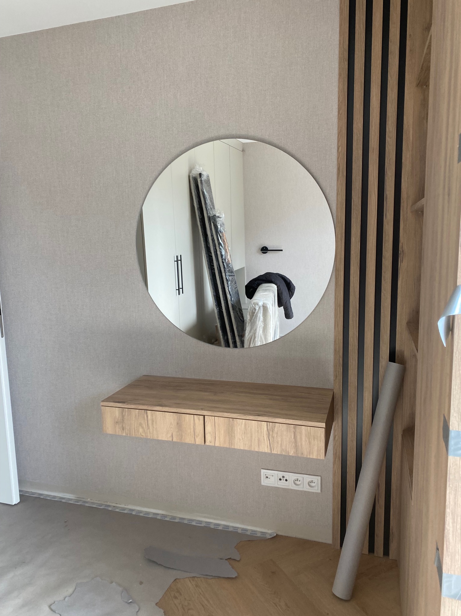 Okrągłe lustro nad drewnianą konsolą z szufladami, odbijające fragment wnętrza z szafą. Dekoracyjne panele ścienne z drewna i czarnych listew. Widoczne gniazdka elektryczne.